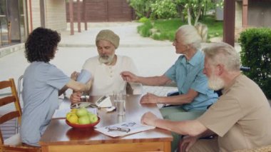 Bahçedeki masada oturan üç erkek ve kadın destekli yaşam tesisi sakininin orta boy görüntüleri. Üniformalı genç hemşire, kol monitörü kullanarak kan basıncını ölçüyor, sohbet ediyor ve gülümsüyor.