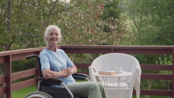 高級退職者コミュニティの庭のテラスで車椅子に座っている移動性障害を持つ幸せな高齢者コーカサス人女性のミディアムフルアークポートレートショット カメラと笑顔を見る — ストック動画