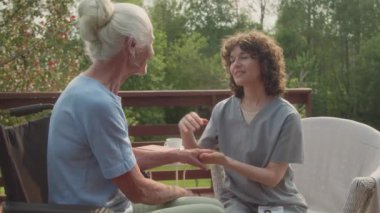 Bahçedeki terasta, tekerlekli sandalyedeki yaşlı bir kadın hastayla oturup sevgi dolu bir konuşma yaparken kolunu tutup okşayan şefkatli, genç bir hemşirenin orta boy görüntüleri.