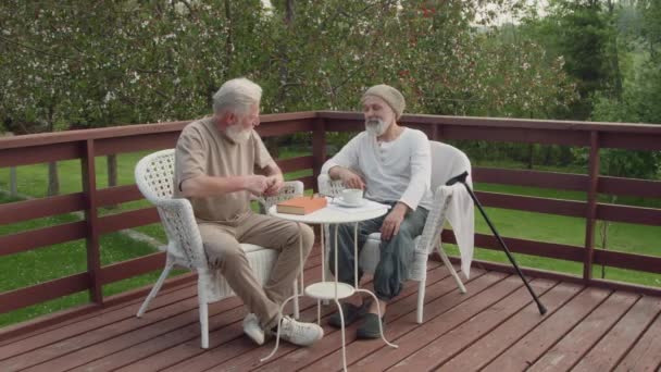 引退した家の庭のアームチェアでリラックスした2人の白人男性住民のフルビデオ チャット ポケットからアルコールとフラスコを取る 友人に与え 試してみることを提供 — ストック動画