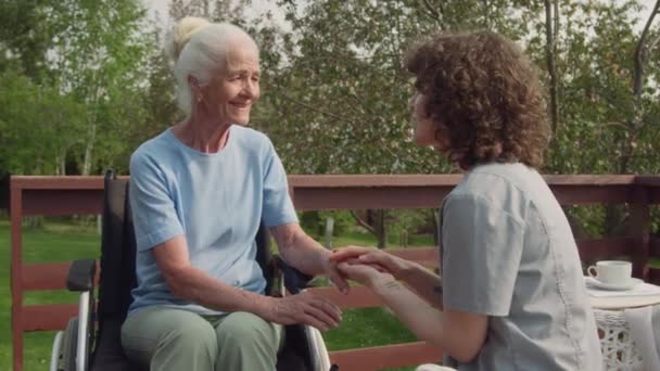 カーリーヘアの若い女性のメディア映像 車椅子で高齢の白人のおばあちゃんとテラスに座っているカジュアルな服 心からの会話をしながら手と肩をストローク — ストック動画