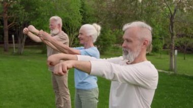 Yaz günü yeşil parkta duran üç yaşlı erkek ve kadının orta ölçekli çekimi. Ekran dışı eğitmene bakıyorlar. Bilek çevirme ve kol esnetme egzersizi yapıyorlar.