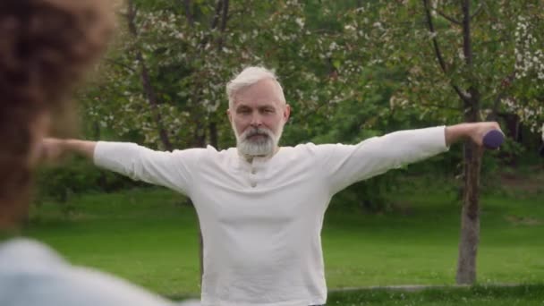 医学療法を受けている間 避難所の庭で灰色の髪とひげで運動する高齢の白人男性の中規模の肩ショット ダンベルで横に上昇 — ストック動画