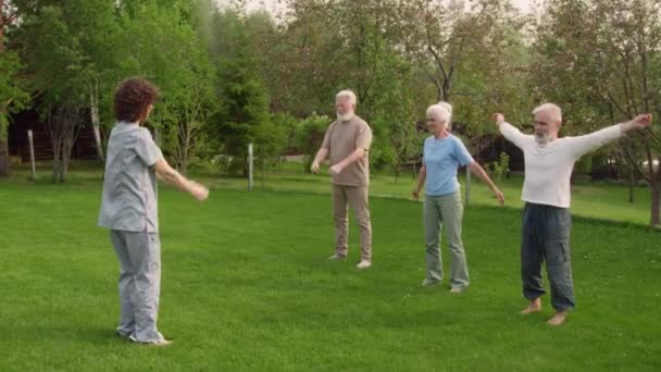 在疗养院的公园或花园 年轻护士站在一群老年男女面前的绿色草坪上 展示着轮臂运动 人们观看着 重复着 — 图库视频影像