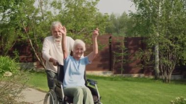 Yaşlı beyaz bir adamın emeklilik bahçesinde koşarken çekilmiş orta boy bir fotoğrafı. Gülen kadın arkadaşıyla tekerlekli sandalye itiyor. O da tezahürat yapıyor ve kollarını sallıyor.