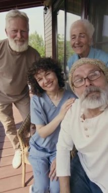 Yaşlı erkek ve kadın Kafkas huzurevi sakinleri ve üniformalı genç hemşire akıllı telefondan eğlenceli video selfie çekerken, el sallarken ve kameraya gülerken orta boy UGC görüntüsü.