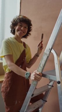 Duvarları merdiven kullanarak boyayan ve evi restore ederken kandıran beyaz bir kadına boya silindiri getiren dikey görüntü.