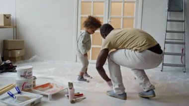 Kirli giysiler içinde Afrika kökenli Amerikalı bir babanın, yere polietilen sürerken ve iki ırklı bir kızla duvar boyamadan önce tüm boya malzemelerini yerleştirirken çekilmiş el kamerası görüntüleri.