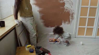 Bekar bir Afrikalı-Amerikalı babanın, melez kızının boya rulosu kullanarak duvarları kahverengiye boyamasını izlerken, yeni dairesinin bakım odasını yenilediğini gösteren bir yan görüntü.