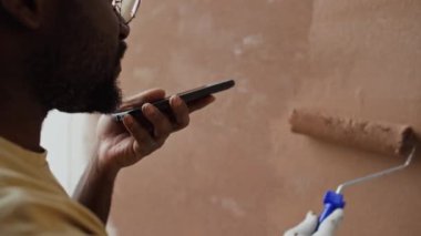 Omuz üstünden, gözlüklü Afrikalı Amerikalı bir adamın hoparlörden akıllı telefon konuşması ve evdeki boya silindiriyle duvarları kahverengiye boyadığı görüntüler.