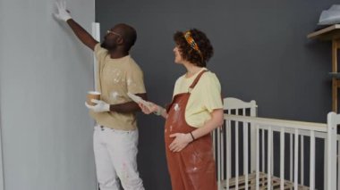 Duvarlar için renk seçen çeşitli ebeveynlerin yan yana olduğu uzun görüntüler. Bebek beşiği yakınlarındaki bebek yuvasında duran renk paletini kullanarak.