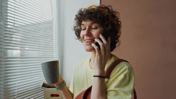 カールヘアとヘアバンドが電話でチャットし 近代的なアパートの窓に立ってコーヒーを飲んで笑顔の白人女性の映像を胸に — ストック動画