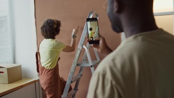黒人の側面映像 彼の最愛のカフカシア人の妻の写真を撮る ポーズし 周囲をだましている間 装飾された部屋で茶色の壁を描く — ストック動画