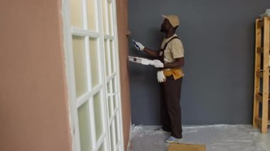 Yan tarafta, çalışan ve yeni apartmanı dekore ederken boya rulosu kullanarak duvarları kahverengiye boyayan siyah bir erkek var.