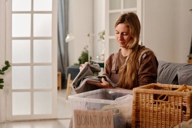 Sarışın kadın gönüllü, eski kıyafetleri katlayıp paketleyip plastik kutuya yerleştirirken bağış merkezi için giysi hazırlıyor.