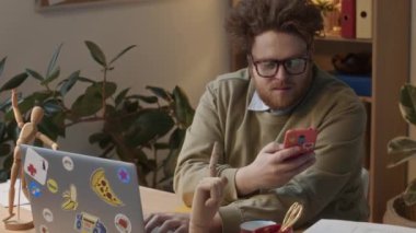 Orta karar kızıl saçlı erkek şirket çalışanının masa başında tahta oyuncaklarla, şirket ofisinde dizüstü bilgisayarla çalışırken, iş saatleri boyunca kişisel konuşmalara cevap verirken görüntüsü.