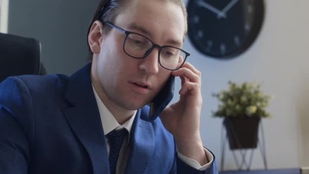 中特写 自恋的男性办公室职员坐在办公桌前 在手机上进行商务对话 看图表 剪掉油光的头发 整理眼镜 — 图库视频影像