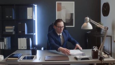 Kafkas erkek şirket çalışanının tam portre görüntüsü. Ofiste oturan gözlüklü, evrak yığınlarını düzelten, kameraya bakan, gülümseyen, sıkıca bağlanmış ellerle poz veren.