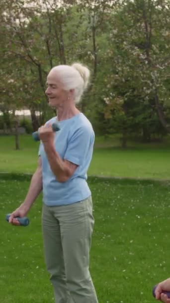 穿着蓝色T恤和裤子站在公园绿色草坪上 用哑铃进行举重训练 同时参加团体健身课的白人老年女士的中 高垂直镜头 — 图库视频影像