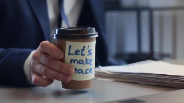 Beyaz takım elbiseli ve gözlüklü beyaz erkek ofis çalışanının yakın çekim görüntüleri. Elinde kağıt yığınıyla masada oturuyor. Uzlaşma notuyla birlikte paket kahve buluyor, gülümsüyor, içiyor ve gülüyor.