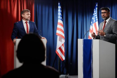 Olgun, ciddi bir erkek politikacı tribünün yanında duruyor ve konferansta konuşma yapan yabancı ülke temsilcisine bakıyor.