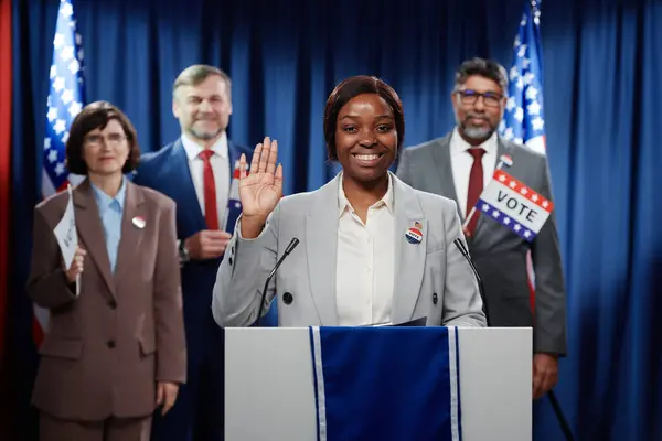 若い陽気な女性政治家が手を振ったり 誓いのジェスチャーを見せたり 代議員に対するプラットフォームで立っている間にカメラを見たりする — ストック写真