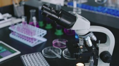 Bilim laboratuarında orta boy mikroskop, mavi ve mor kimyasal çözeltili şişeler, maddeli petri kapları, raflarda sıra sıra test tüpleri,