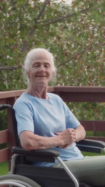 高級退職者のコミュニティの庭のテラスで車椅子に座って移動性の障害を持つ幸せな高齢者コーカサス人の女性の中型垂直の肖像画は カメラと笑顔を見ています — ストック動画