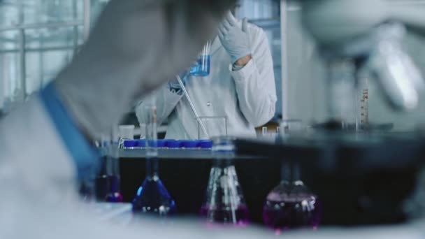 中国女性研究人员在玻璃瓶中嗅出蓝色溶液的中照 在化学实验中 同事们对显微镜的前景进行了模糊的观察 — 图库视频影像