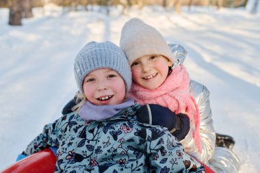 İki sevimli, dişlek gülümsemeli kız kameraya bakarken bir tanesi sevimli arkadaşının önünde kar tüpünün üzerinde oturuyor.