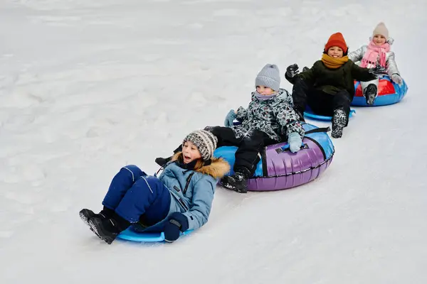 一排排穿着冬衣的可爱的快乐孩子坐在滑雪板和雪管上 骑着马在自然环境中下山 免版税图库图片