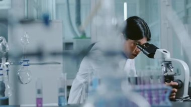 Çinli kadın araştırma kimya laboratuarı çalışanının mikroskop altında numuneleri incelerken, sonra dizüstü bilgisayarda yazarken, deney sırasında gözlemleri kaydederken orta kavisli bir çekim.