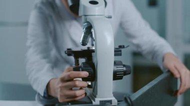 Beyaz önlüklü Asyalı kadın bilim laboratuvar çalışanı mikroskoba bakarken ve düğmeyi odaklamak için çevirirken yakın çekim.