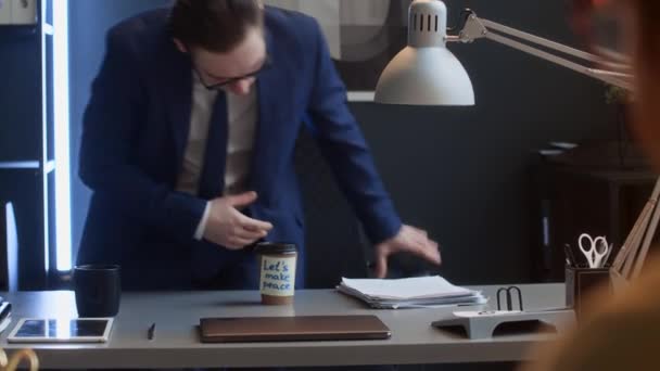 身着西服 戴着眼镜 拿着大堆文件走到办公桌前的白人男性公司员工的中景照片 照片中 他们看到一只带着和平字条的外卖咖啡杯 面带微笑 大显身手 — 图库视频影像