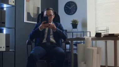 İş adamı kılığında, kravatlı ve gözlüklü bir adamın akıllı telefon kullanarak sandalyede oturması, sonra da bir yığın kağıtla yanımızdan geçip zalimce gülmesi.