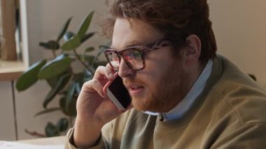 Yakın çekim, şapşal kızıl saçlı, gözlüklü, masasında oturan, cep telefonuyla konuşan, el kol hareketleriyle işaret eden, ahşap oyuncak elli beyaz erkek ofis çalışanı.