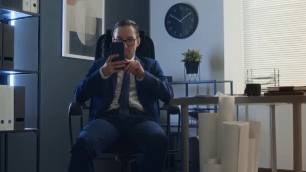 穿着西服 打着领带 戴着眼镜 坐在办公桌前 用智能手机 拿着大堆文件走在同事身边 恶狠狠地笑着的职场小伙拍了照 — 图库视频影像