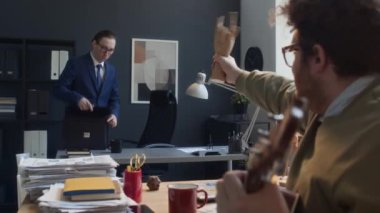 Orta boy, kızıl saçlı, beyaz bir adamın sabah ofiste oturup, iş için gelen iş arkadaşını taciz ederken, ahşap ellerle ona el sallarken görüntüsü.