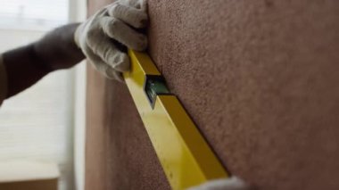 Odanın yenilenmesi sırasında ruhsal seviyeyi kullanarak kahverengi duvara işaret yapan isimsiz tamirci eldivenleri giymiş ellerin yan görüntüsü.