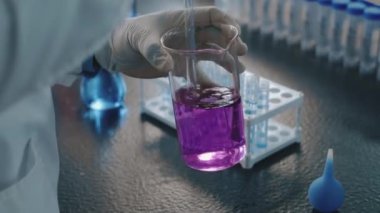 Beyaz önlüklü ve eldivenli tanımlanamayan kadın laboratuar teknisyeni veya kimyagerin elleri, cam samanla hafif mor permanganat solüsyonunu karıştırırken, testlere hazırlanıyor.