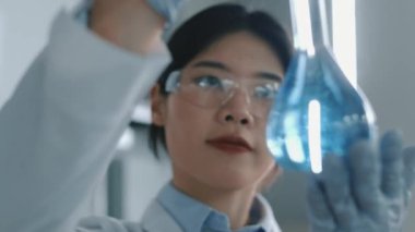 Gözlüklü, beyaz önlüklü ve eldivenli genç Çinli kadın araştırmacının mavi kimyasal çözeltili cam şişeyle görsel özellikleri incelerken yakın plan çekimleri.