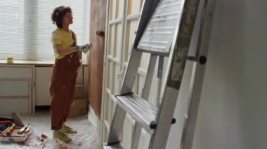 Bebek odasında duvardan duvara A şeklindeki merdivenin yan görüntüleri. Beyaz hamile bir kadın arka planda kahverengi paten ile duvarları boyuyor.