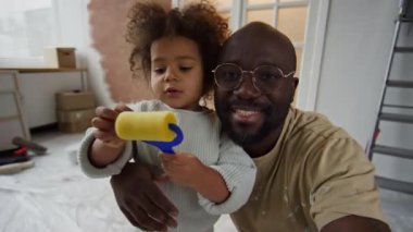 POV UGC, gözlüklü Afro-Amerikan bir adamın video görüntüleri. Küçük melez kızıyla video blogu için kayıt yapıyor. Elinde mini boya silindiri tutuyor ve oda yenileme sırasında kameraya bakıyor.