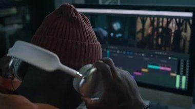 Örgü şapka takan, kulaklık takan, profesyonel grafik tasarım yazılımında film görüntülerini düzenlemeye hazırlanan Afrikalı Amerikalı bir adamın yakın çekim arka plan odağı.