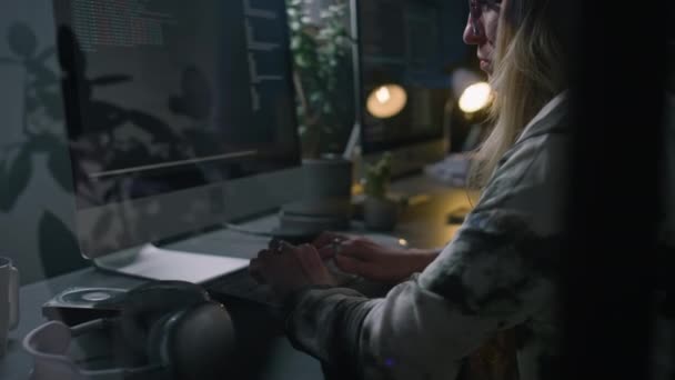 通过白种人女软件开发商Beanie的玻璃墙 夜间坐在黑暗公司办公室桌子前的眼镜 键盘打字 为项目编写计算机代码的中等特写镜头 — 图库视频影像