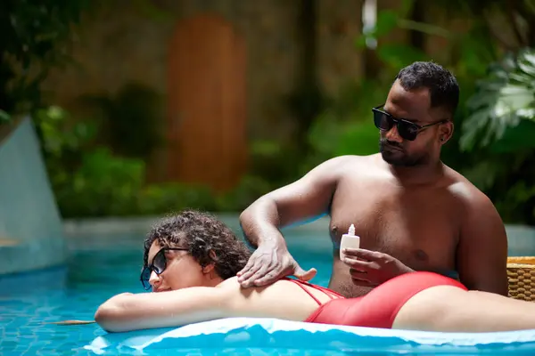 若いセレンカップルのイディエリックなシーンは 青い水とプールで時間を過ごしながら 男は少女の背中に日焼け止めのローションを適用しています ストック写真