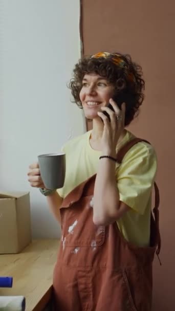 改装された家の窓で立っているコーヒーまたは茶休憩中に電話会話を持つジャンプスーツの陽気な白人女性の垂直側の映像 — ストック動画