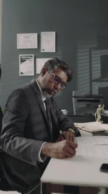 Gözlüklü ve takım elbiseli çift ırklı avukatın adli yardım bürosunda çalışırken kağıt işleriyle uğraşırken çekilmiş dikey görüntüler.