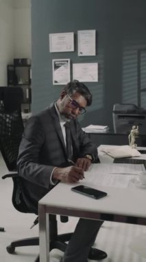 Pahalı takım elbiseli ve gözlüklü ciddi bir çift ırklı avukatın modern hukuk bürosundaki masada belgelerle çalışırken çekilmiş dikey görüntüler.