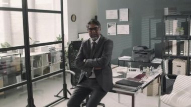 Masada oturan ve modern ofiste kollarını kavuşturarak kameraya poz veren kendinden emin çift ırklı avukatın orta boy portresi.